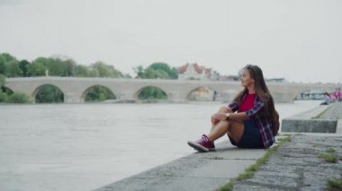 Spor ayakkabılı şık kız Regen Nehri kıyısında oturuyor, Regensburg Stone Köprüsü.