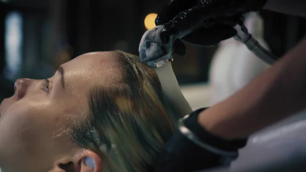 Hairdresser in black gloves carefully washes females hair with conditioner in sink in hair salon, and then turns off the water. Closeup