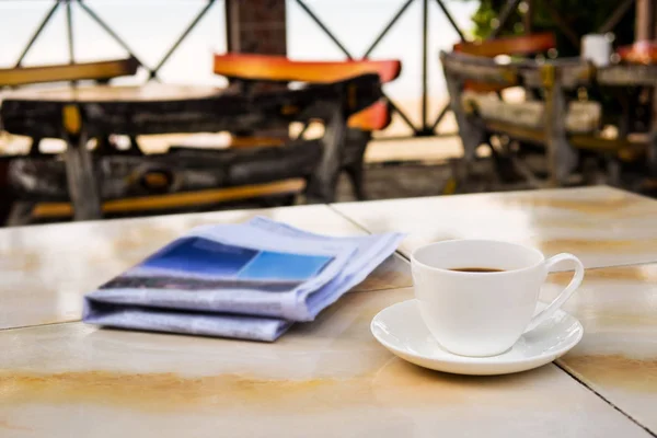 Feche a xícara de café branco na mesa amarela na praia de areia do nascer do sol com jornal pela manhã — Fotografia de Stock