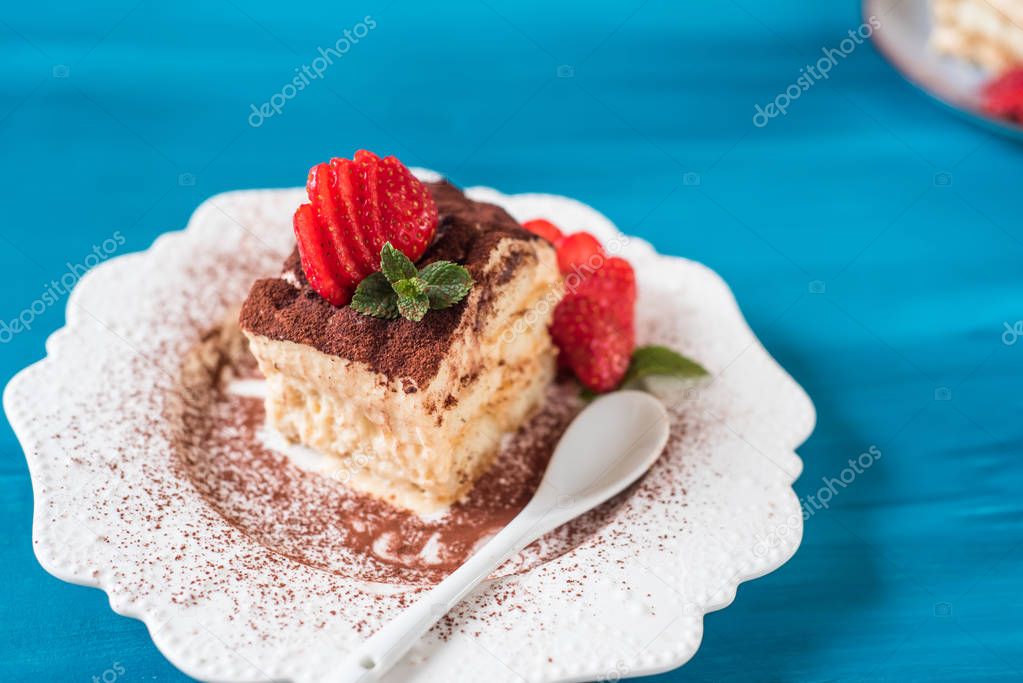 Piece of tiramisu cake with mint and strawberries on a blue background