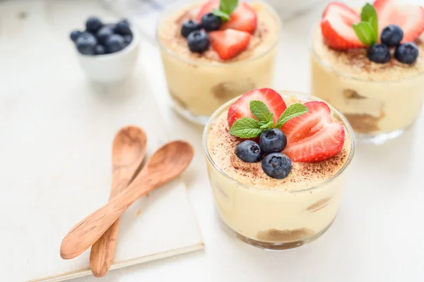 Hecho en casa, exquisito postre tiramisú en vasos decorados con fresa, arándano, menta sobre mesa de madera blanca — Foto de Stock