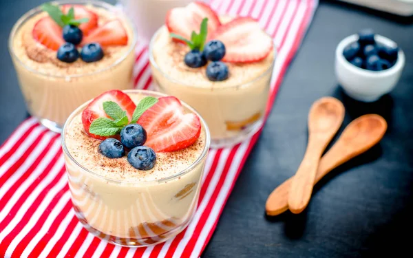 Fatto in casa, squisito dessert tiramisù in bicchieri decorati con fragola, mirtillo, menta su tavolo di legno nero — Foto Stock