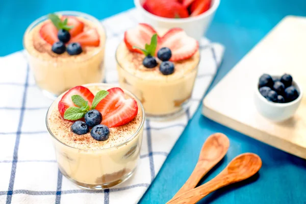 Homemade, exquisite dessert tiramisu in glasses decorated with strawberry, blueberry, mint on blue wooden table — Stock Photo, Image