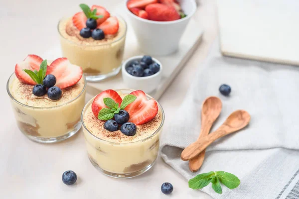 Homemade, exquisite dessert tiramisu in glasses decorated with strawberry, blueberry, mint on white wooden table — Stock Photo, Image