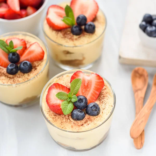 Homemade, exquisite dessert tiramisu in glasses decorated with strawberry, blueberry, mint on white wooden table, top view. — Stock Photo, Image