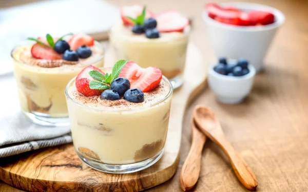 Homemade, exquisite dessert tiramisu in glasses decorated with strawberry, blueberry, mint on white wooden table, top view. — Stock Photo, Image