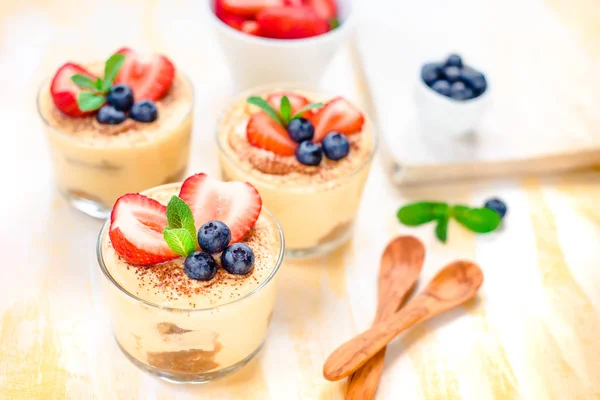 Homemade, exquisite dessert tiramisu in glasses decorated with strawberry, blueberry, mint on white wooden table, top view. — Stock Photo, Image