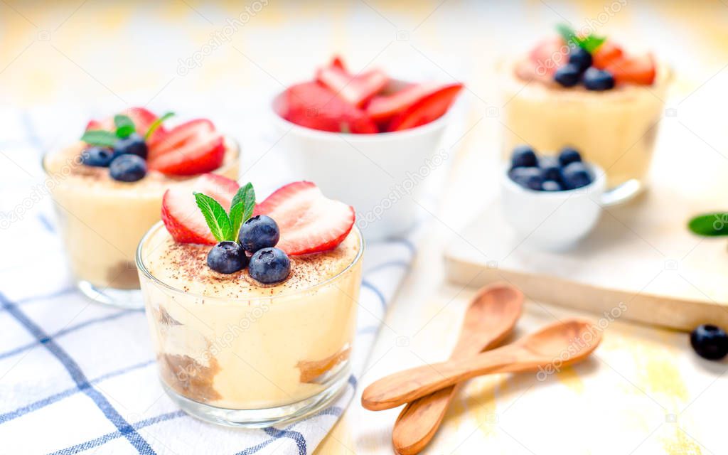 homemade, exquisite dessert tiramisu in glasses decorated with strawberry, blueberry, mint on white wooden table