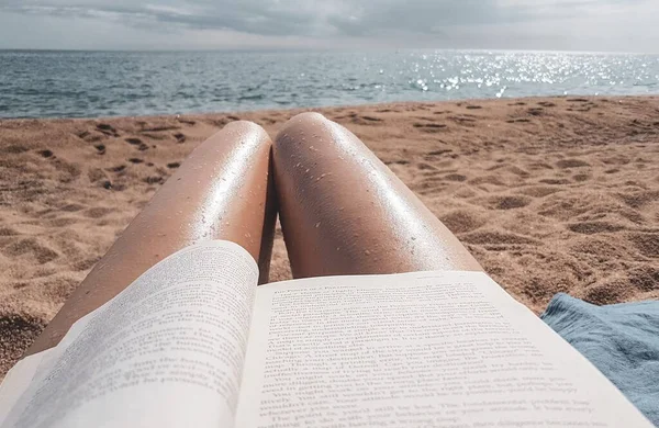 Una giovane ragazza legge un libro sulla spiaggia durante una vacanza. Sexy gambe nude, libro, sabbia, spiaggia, mare . — Foto Stock