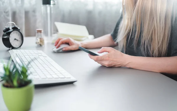 Νεαρή γυναίκα, ένας online έμπορος κάθεται σε μια επιφάνεια εργασίας και λειτουργεί μέσω ενός smartphone — Φωτογραφία Αρχείου