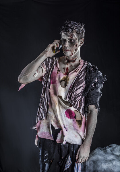 Male zombie using cell phone calling someone, standing on black smoky background, half body shot