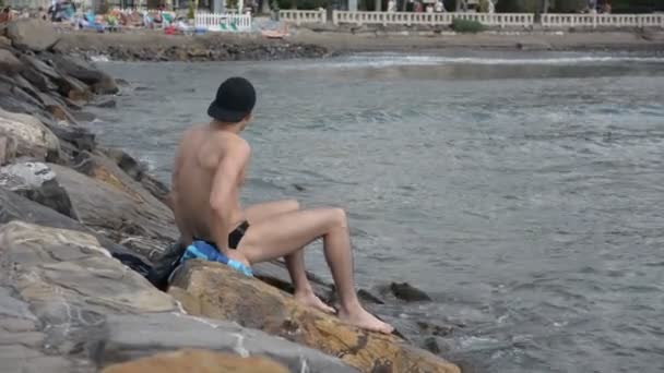Jonge shirtless atletische man zit op rots door zee — Stockvideo