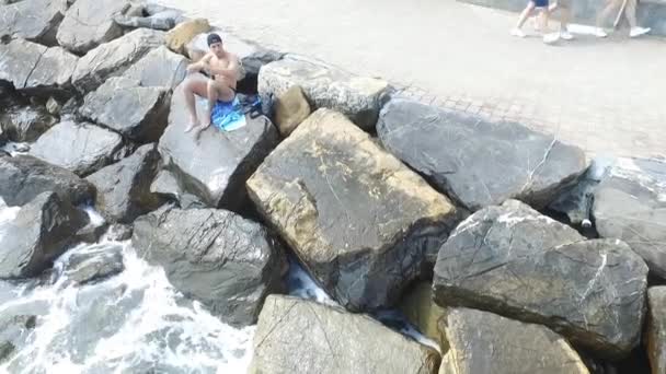 Молодой спортсмен без рубашки сидит на скале у моря — стоковое видео