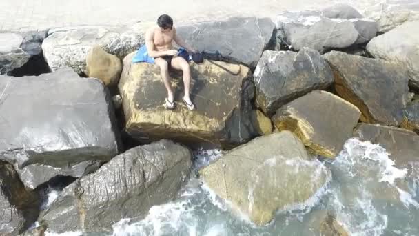 Νεαρός shirtless αθλητική άνδρας κάθεται στο βράχο από την θάλασσα — Αρχείο Βίντεο