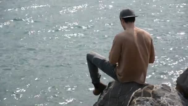 Молодой человек сидит и думает перед морем — стоковое видео