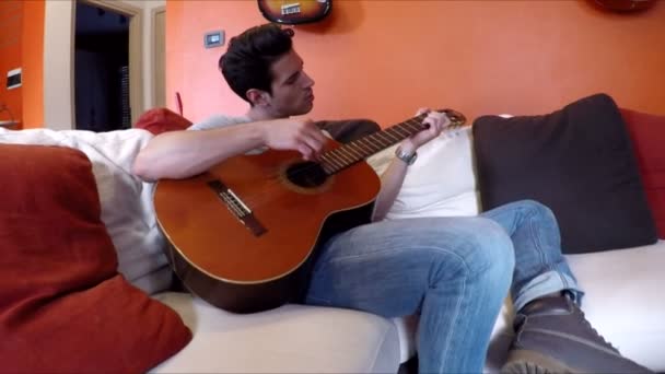 Joven en el sofá tocando la guitarra en casa — Vídeos de Stock