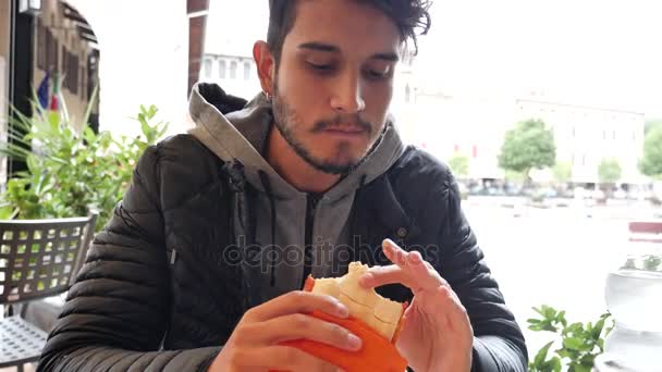 カフェに座っている間にサンドイッチを持っている若い男 — ストック動画