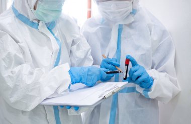 Korunma giysisi giymiş Asyalı kadın doktor karantina hasta dosyasında maske yazıyor. Koronavirüs taraması için kan örneğiyle birlikte test tüpü tutuyor. Coronavirus, covid-19 konsepti.