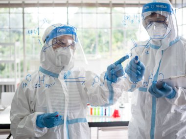 Laboratuvarda PPE giysisi ve yüz maskesi olan bilim insanları ve mikrobiyologlar koronavirüs enfeksiyonu için aşı veya ilaç üretmek için kimyasal formül hesaplarlar. Covid-19, laboratuvar ve aşı konsepti..