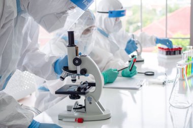PPE giysisi ve yüz maskesi olan bilim insanları ve mikrobiyologlar laboratuarda test tüpü ve mikroskop tutuyor, koronavirüs enfeksiyonu için aşı ya da tedavi buluyorlar. Covid-19, laboratuvar ve aşı konsepti..