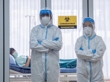 İki Asyalı doktor N95 maskeli ve yüz kalkanlı PPE kıyafeti giyer, koronavirüs enfeksiyonlu hastayı negatif basınç odasında tedavi eder, karantina uyarısı alan işareti ile etiketlenir. Coronavirus ve covid19 konsepti.