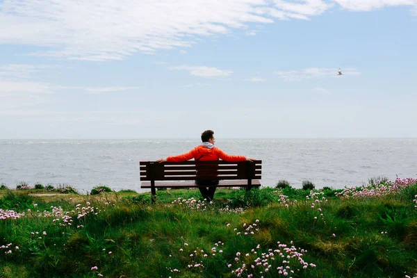 Молодой человек сидит на скамейке и смотрит на море — стоковое фото