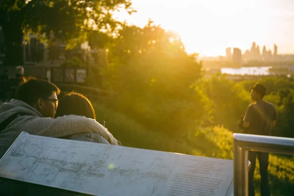 Cuplu așezat pe pământ pe vârful dealului de la Greenwich Park, Londra, îmbrățișându-se și urmărind apusul soarelui — Fotografie, imagine de stoc