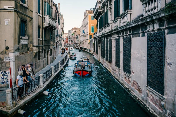 Venetian street canal with various boats on it, tourists walk alongside. — Stock Photo, Image
