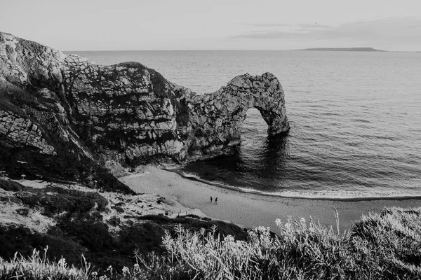 Black & white view from the hill over people walking on a beach by the sea and Durdle Door, a natural limestone arch on Dorset\'s Jurassic Coastline, UK.