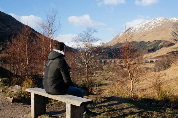 A man sits on a wooden bench, looking at Glenfinnan viaduct, Scotland, on a sunny spring day.