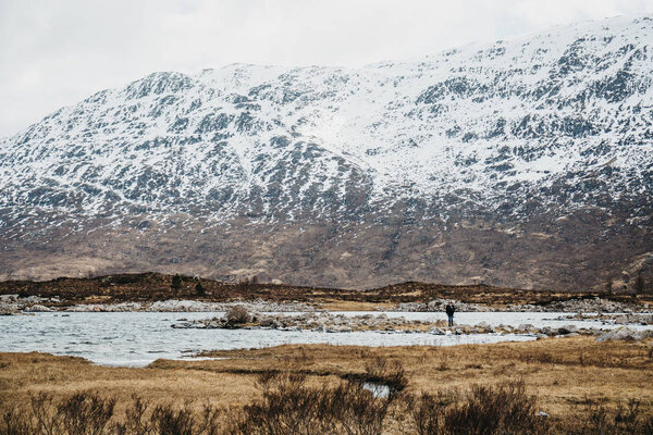 Man walking by scowcapped mountains and loch in Scottish Highlands, Scotland, in spring. 