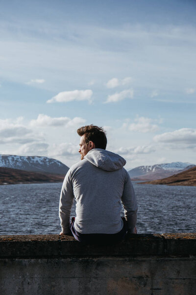 A young handsome man sits on the edge of the water on a sunny day, looking the side, snow-capped mountains, sky and clouds in the background.