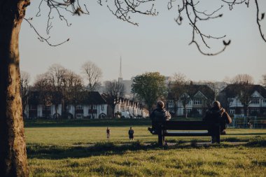 Londra, İngiltere - 22 Mart 2020: Bankın karşı tarafında oturan iki adamın Broomfield Park 'ta, Londra Enfield Borough' daki Palmers Green 'deki halk parkında top oynayan insanları izlemesi.