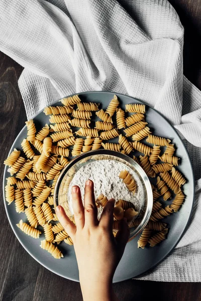 Ahşap arka planda çiğ makarna, üst manzara. Beyaz havlunun üzerinde un olan bir tabakta kurutulmuş makarna. Çocuğun eli makarnaya uzanır..