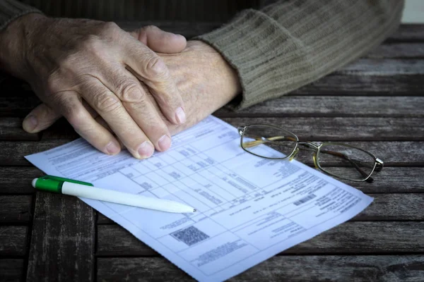 Hands of old senior man fills in utility bills on wooden table. Planning month budget, calculating expen. Wrinkled palm of close up, copy space