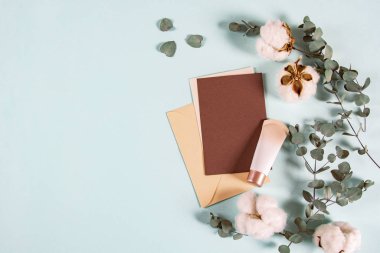 Boş kâğıt zarflar, davetiye mektupları, okaliptüs yapraklı mektuplar, mavi arka planda krema kavanozu ve pamuk çiçekleri, düz yatak, üst manzara. Doğal organik güzellik ürünü