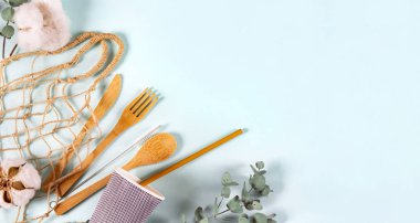 Eko doğal ahşap kaşık, çatal, bıçak, tabak, kağıt bardak, ip torbası ağı, okaliptüs yaprakları ve arka planda pamuk çiçekleri, afiş. Sıfır atık, plastiksiz konsept. Düz konum, üst görünüm, kopyalama alanı