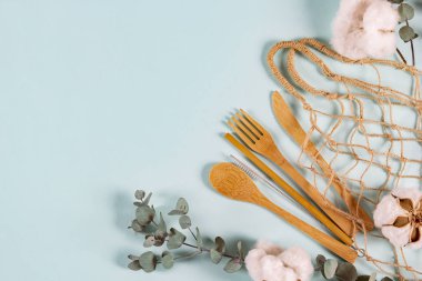 Çevreye özgü ahşap bambu çatal bıçak seti, tel torba örgü, okaliptüs yaprakları ve pastel mavi arka planda pamuk çiçekleri. Sıfır atık, plastiksiz konsept. Düz konum, üst görünüm, kopyalama alanı