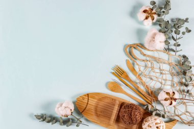 Eko doğal ahşap kaşık, çatal, bıçak, tabak, ip torbası örgüsü, okaliptüs yaprakları ve pastel mavi arka planda pamuk çiçekleri. Sıfır atık, plastiksiz konsept. Düz konum, üst görünüm, kopyalama alanı