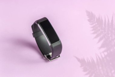 Açık pembe arka planda akıllı takip cihazı. Eğrelti otu gölgesi ve spor bileziği olan siyah fitness saati.