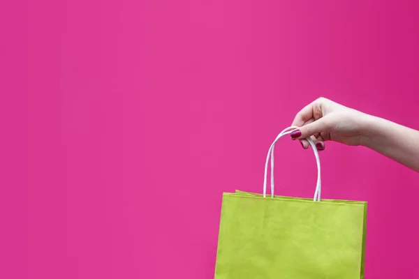 Bright green paper bag in woman hand on pink fuchsia background. Concept of shopping, delivery of goods, purchases.