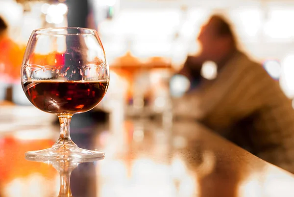 Glass of cognac on a bar stand with a blurred silhouette of a drinking man