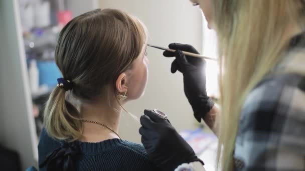 A maquilhadora desenhando sobrancelhas para uma garota bonita com um pincel profissional na frente de um espelho em um salão de beleza . — Vídeo de Stock