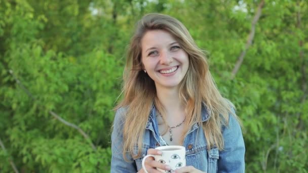 Cute girl with long hair laughing with a cup in hands against the background of green bushes. Pretty blond girl in jeans jacket smiles in a slow motion. — Stock Video