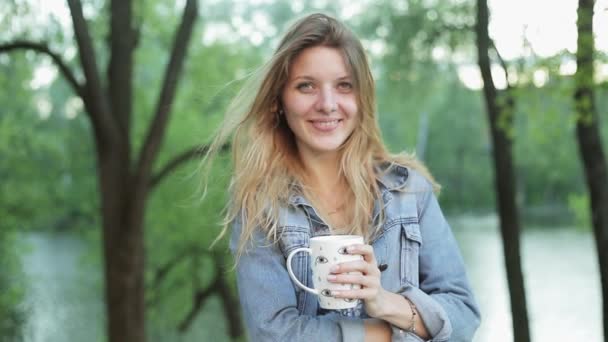 Šťastná dívka s dlouhými vlasy drží šálek čaje nebo kávy v rukou na pozadí rozmazané přírody. Hezká žena v jeans bunda úsměvy a pití nápoj. Zpomalený záběr. — Stock video
