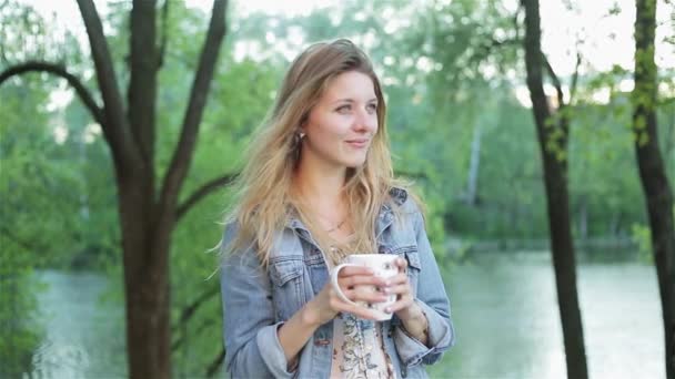 Happy girl with long hair holds a cup of tea or coffee in hands against the blurred nature background. Pretty woman in jeans jacket smiles and drinks beverage. Slow-motion shot. — Stock Video