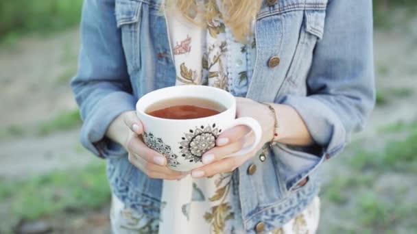 Menina com cabelos longos segura uma xícara de chá em suas mãos contra o fundo da natureza borrada. Mulher bonita em jeans jaqueta segura uma xícara com bebida . — Vídeo de Stock
