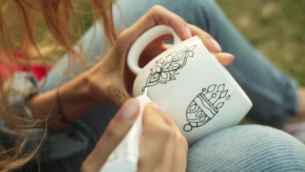 Primer plano de las manos femeninas dibujando un patrón en una taza blanca. Chica pinta adorno sentado en la hierba en la noche . — Vídeos de Stock