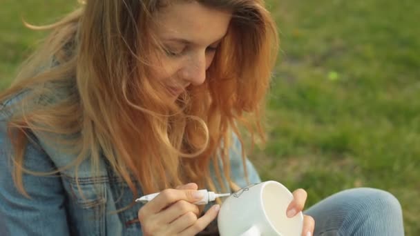 Närbild av kvinnliga händer rita ett mönster med färg från en tub på en vit mugg. Leende flicka målar prydnad sitter på gräset på kvällen. — Stockvideo