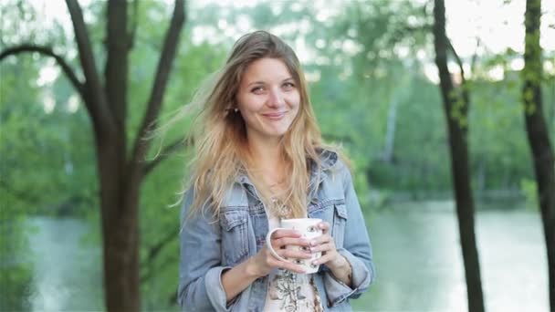 Young shy girl with long hair holds a cup of tea or coffee in hands against the blurred nature background. Pretty woman in jeans jacket smiles and drinks beverage. — Stock Video